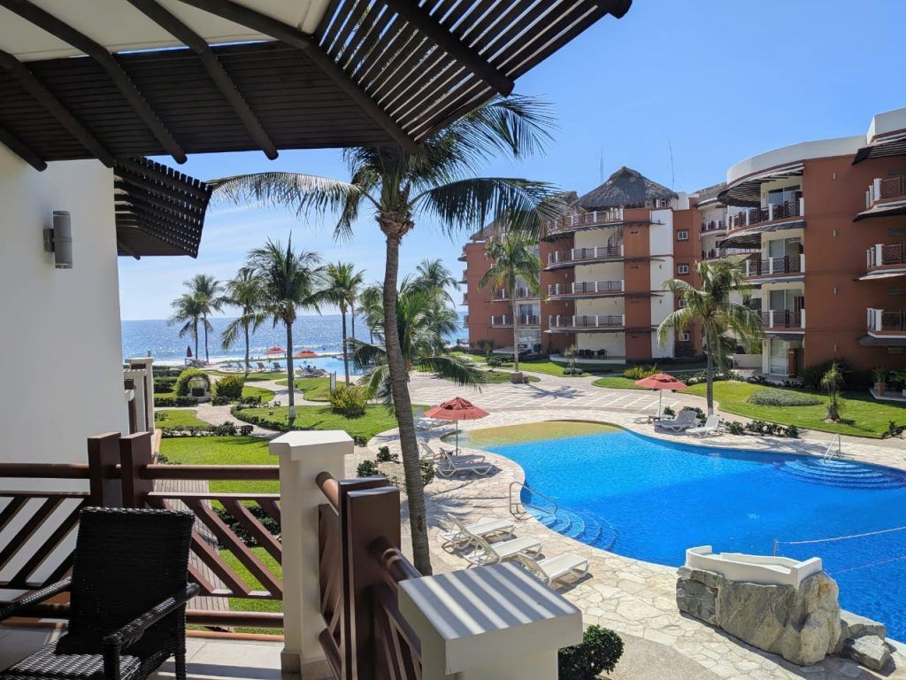 View from a balcony overlooking a pool, resort buildings, palm trees, and the ocean. Bright sunlight.