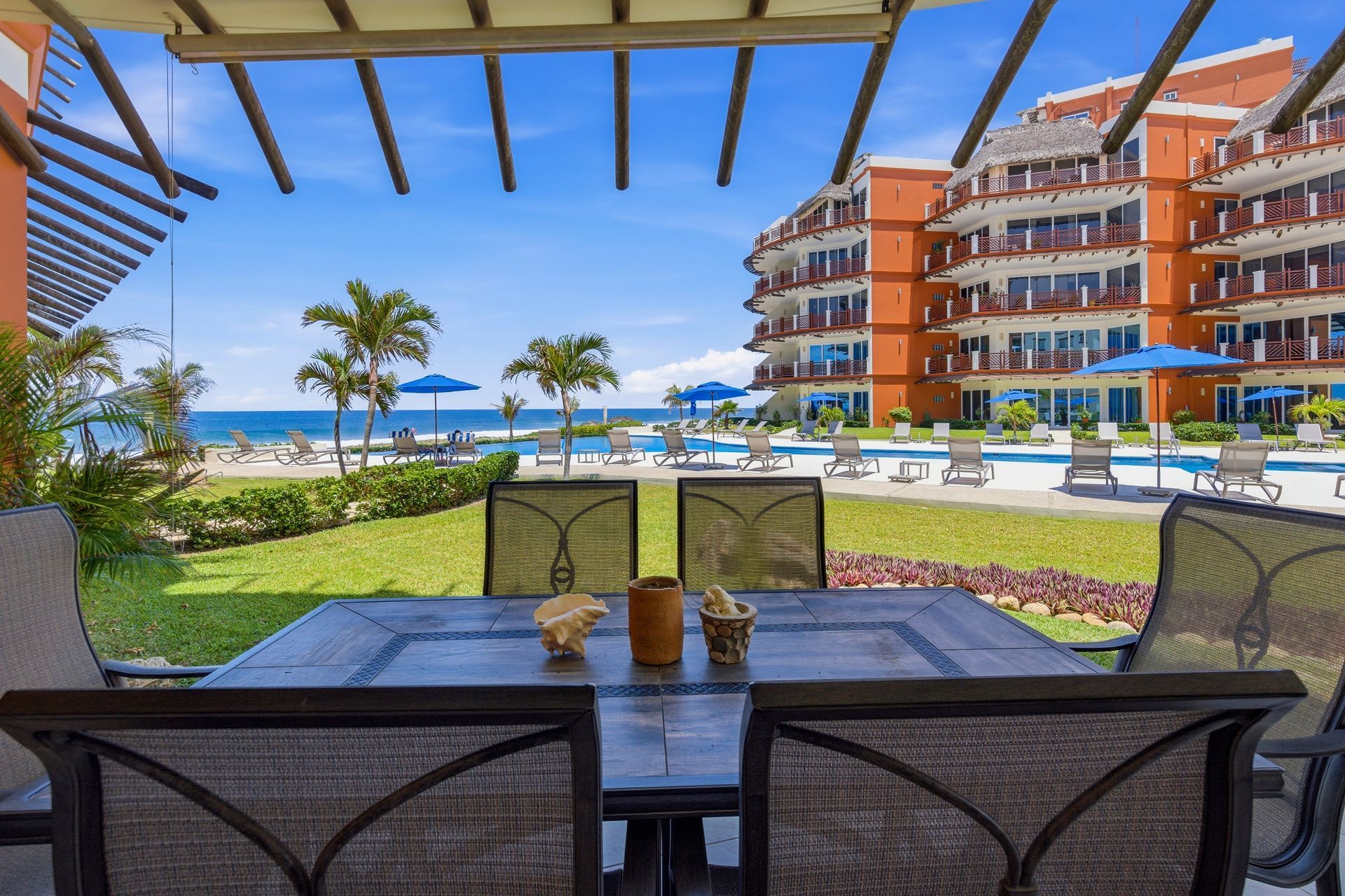 Shaded patio dining table overlooking a resort lawn, pool, palm trees, and ocean