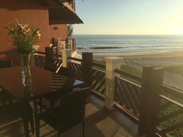 Balcony overlooking a beach at sunset. Table with flowers, railing, and ocean visible.