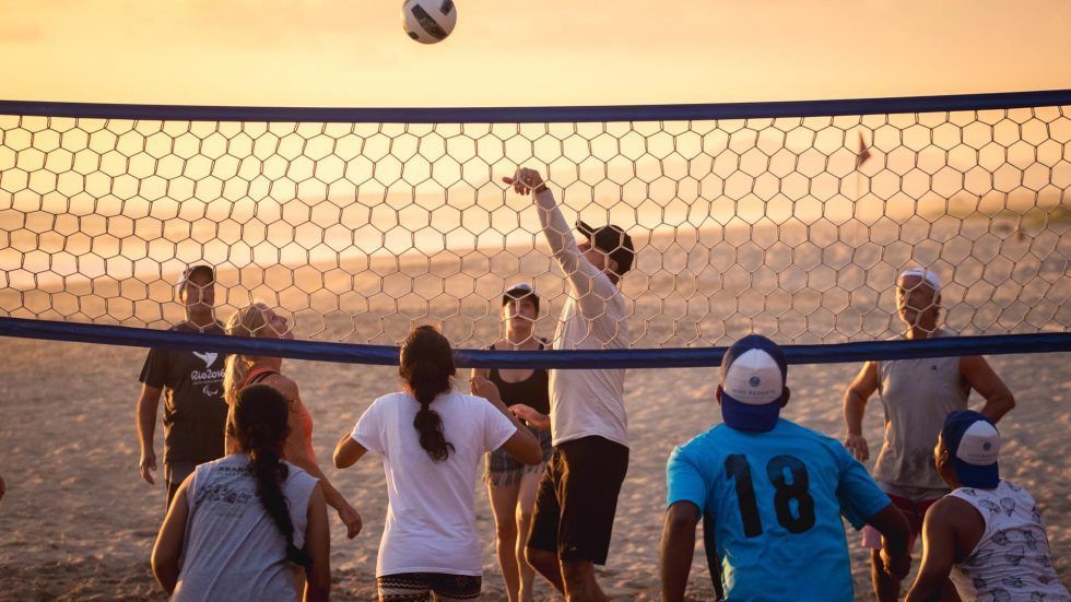 Volleyball players jump at the net on a beach at sunset, with one reaching to block the ball.