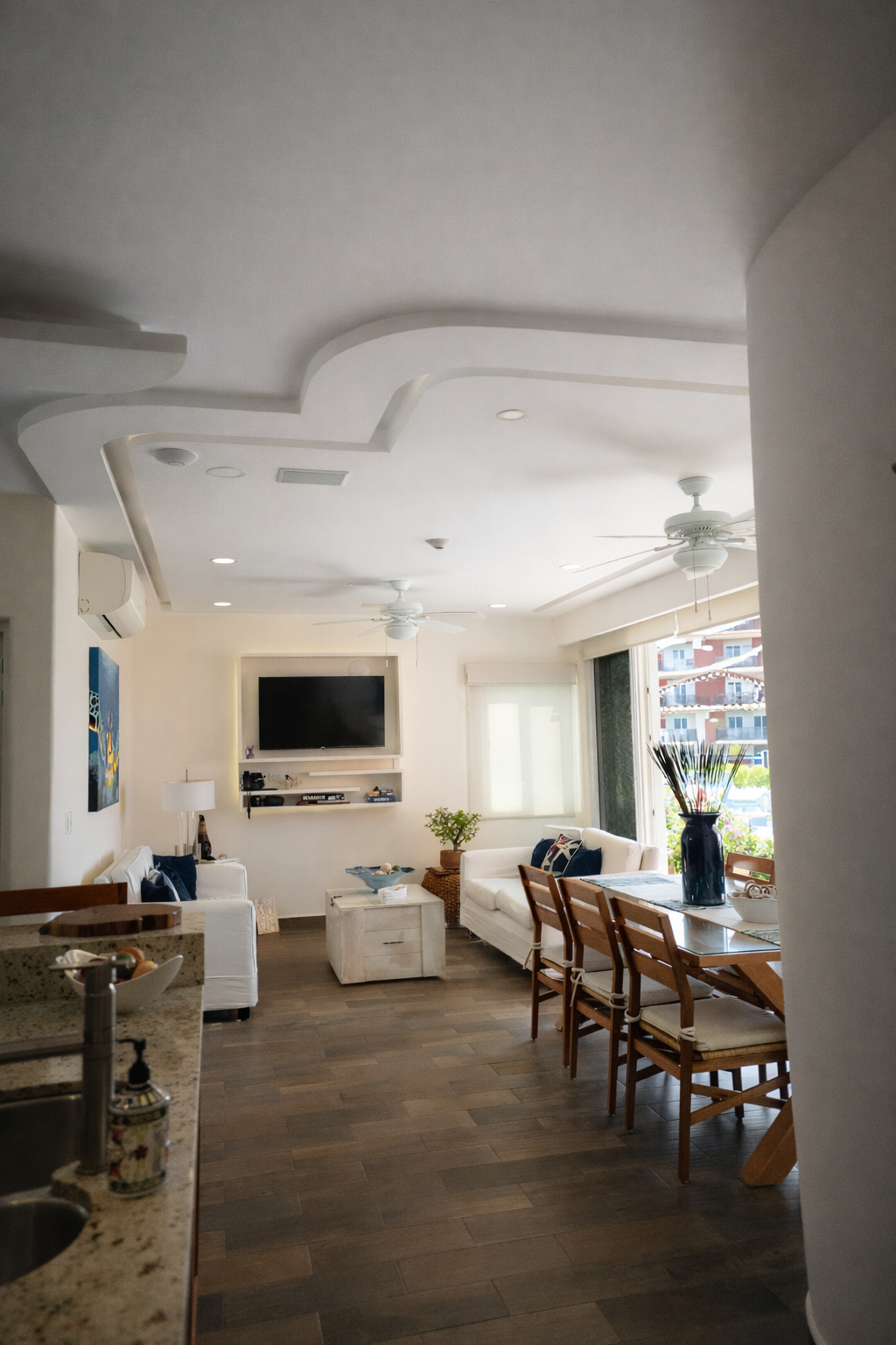Bright living room with wood floors, white ceiling, dining table, sofa, TV, and large windows.