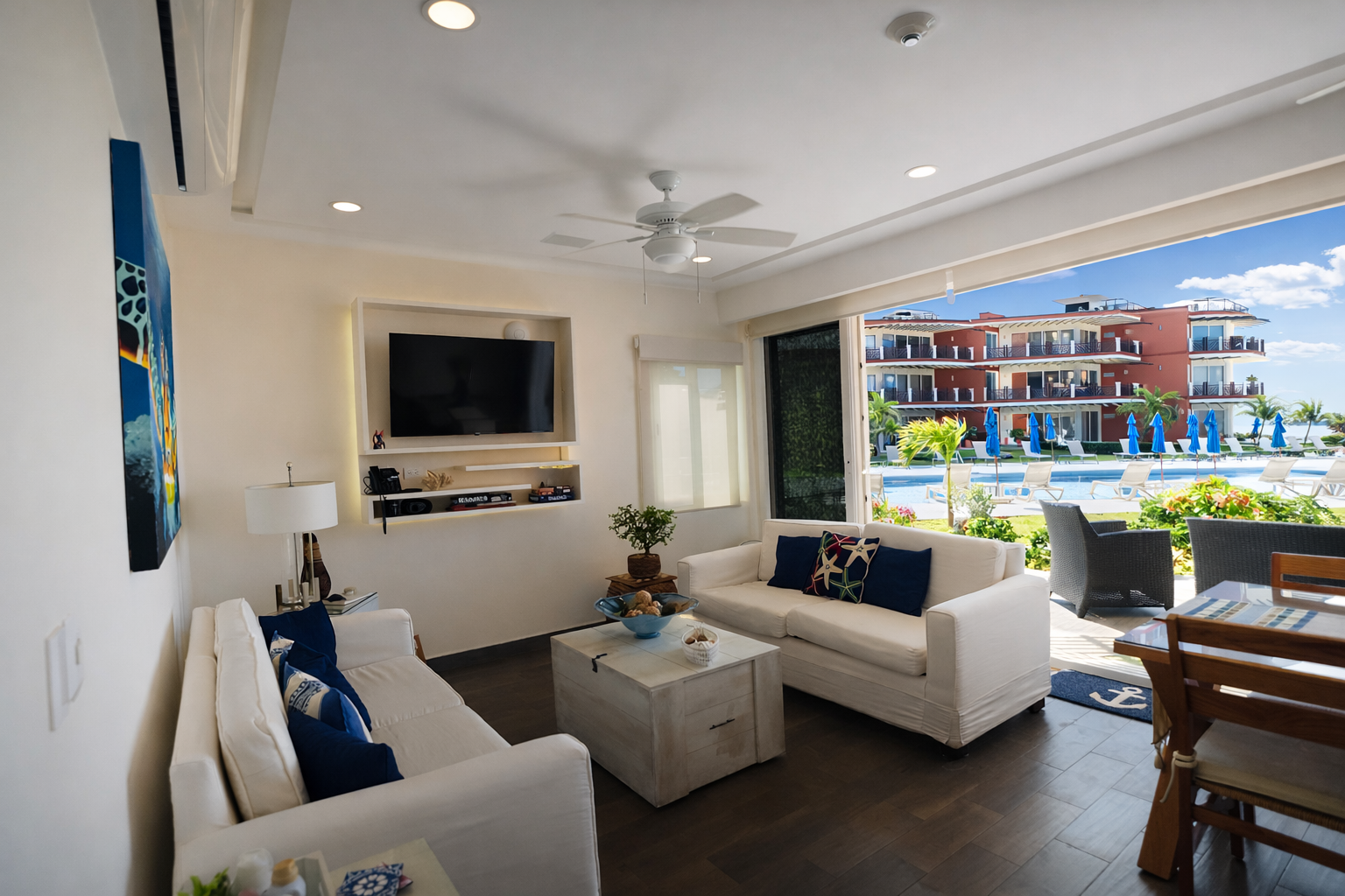 Bright modern living room with white sofas, wall TV, and open view to a sunny balcony and pool.