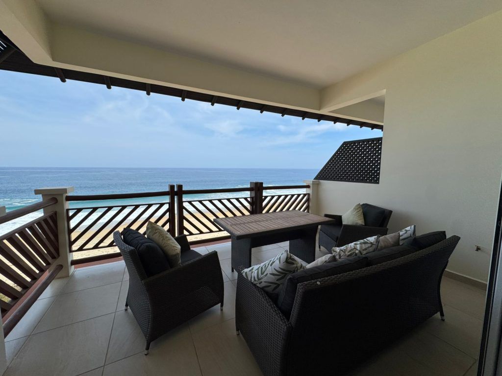 Patio with ocean view; black wicker furniture, wooden railing, blue sky and water.