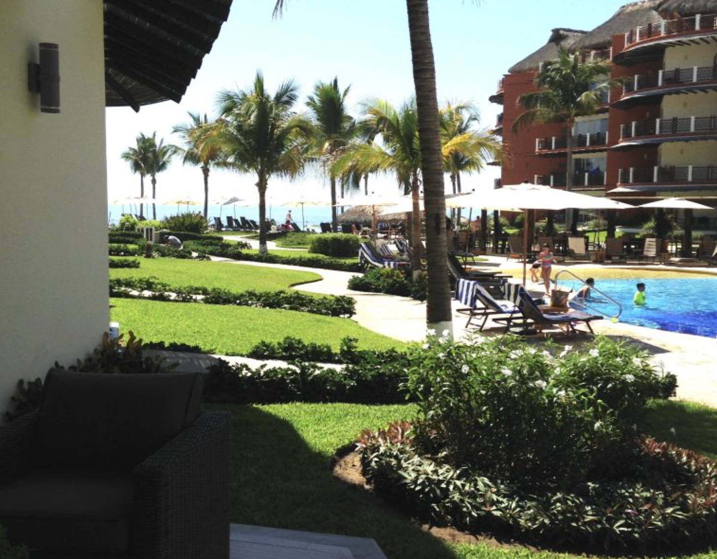 Lush resort grounds with pool, ocean view, palm trees, and a building on a sunny day.