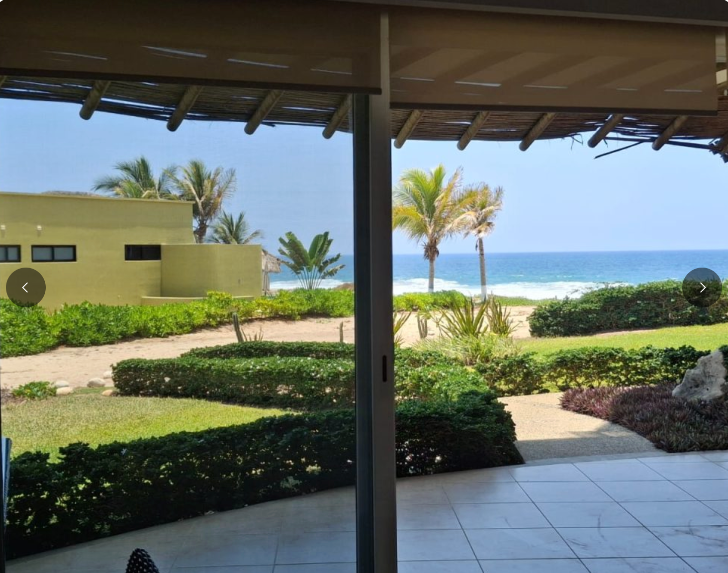 View of a beach and ocean from a patio, lush green landscaping, blue water and sky.