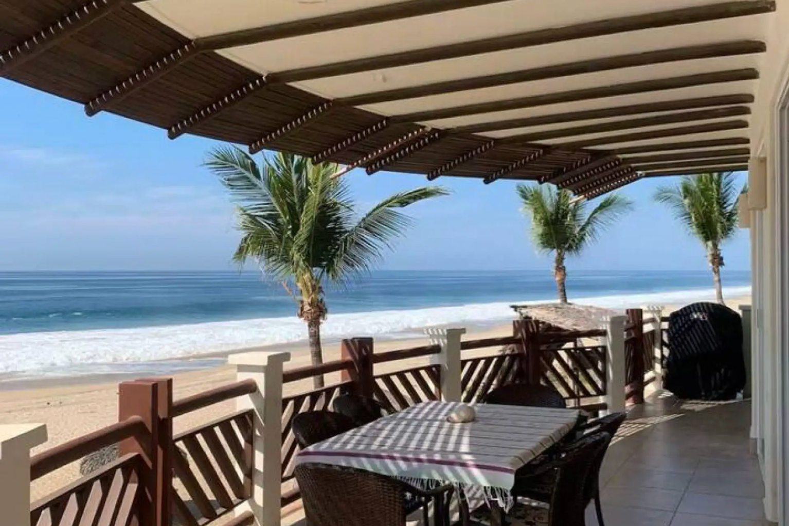 Beachfront patio with table, chairs, and ocean view under a shaded pergola.