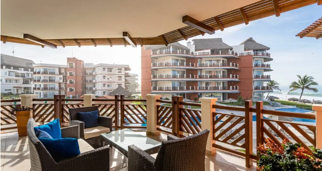 Balcony with wicker chairs overlooking a resort pool and ocean, framed by wooden railing and canopy.