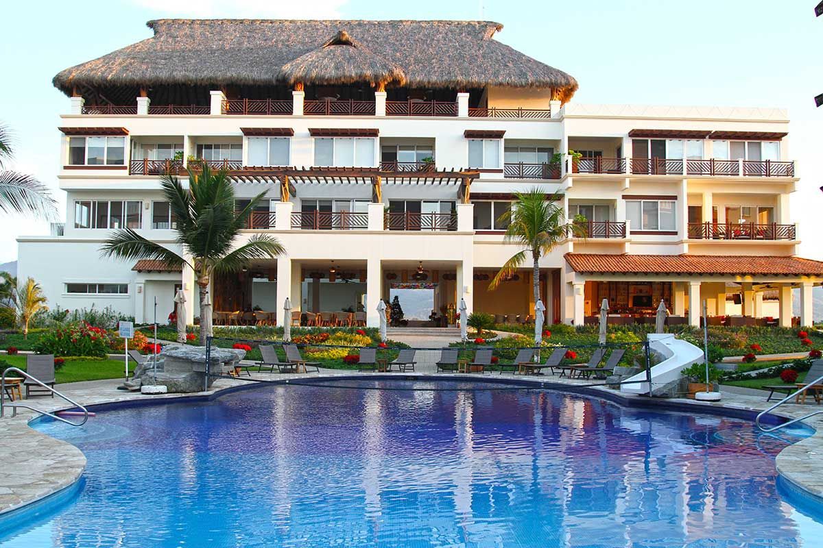 Hotel with a thatched roof, balconies, and a blue pool in front.