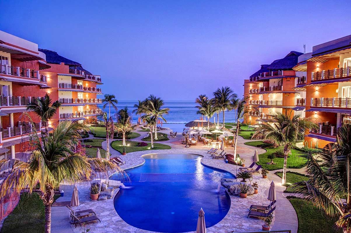 Resort pool at dusk, with buildings, palm trees, and beach in the background.