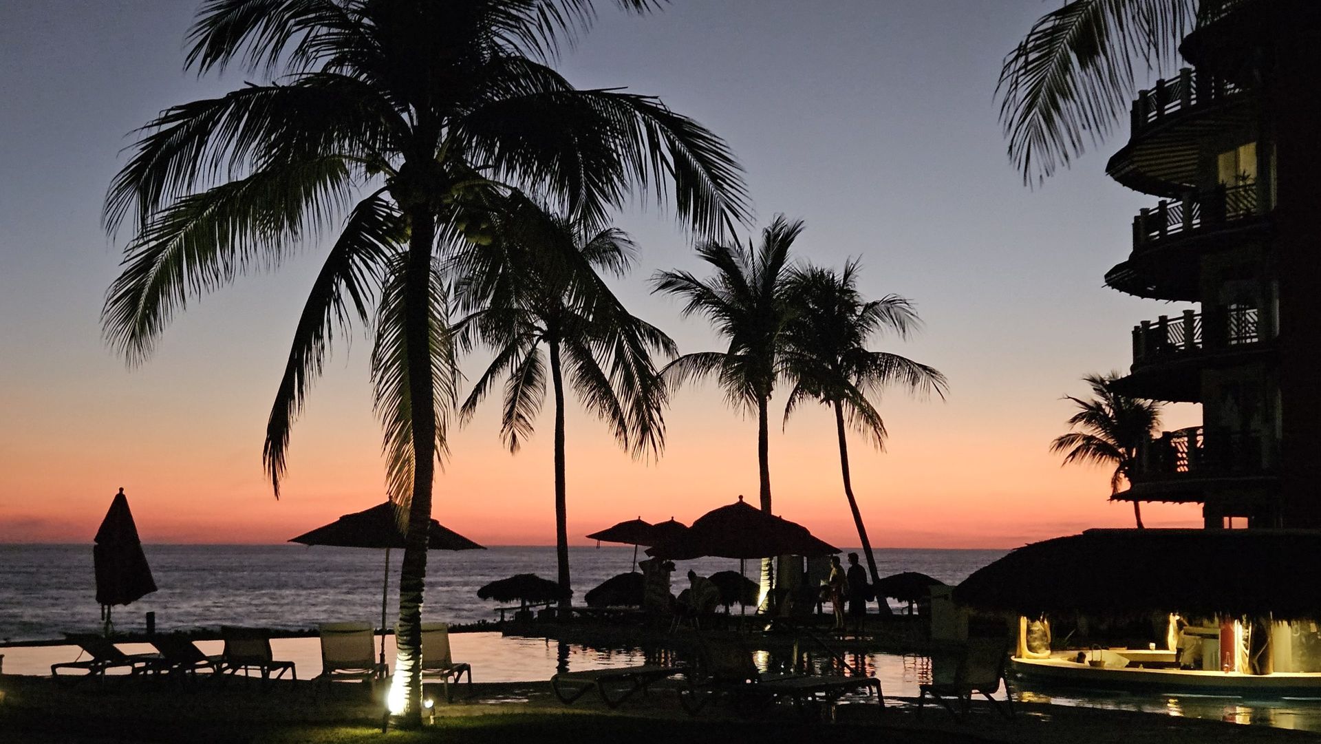 Palm trees silhouetted against a colorful sunset over a beach resort.