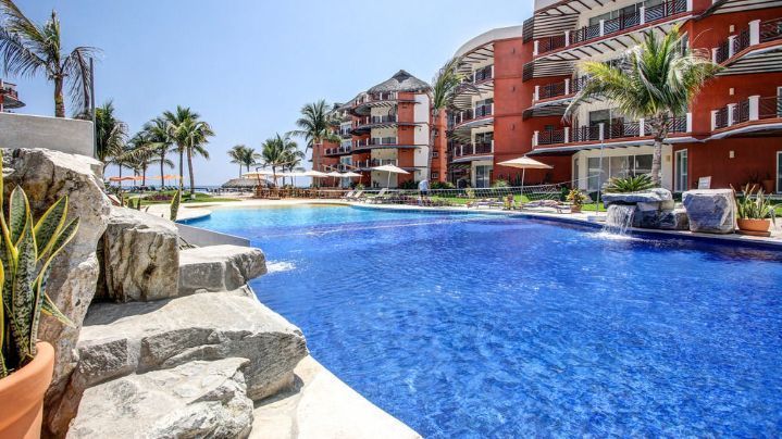 Pool with blue tile near a building with red and white balconies, palm trees, and ocean views.