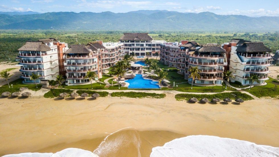 Beachfront resort with multiple buildings, pool, and ocean view; mountains in the background.