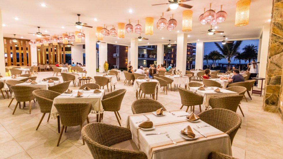 Restaurant interior with tables, chairs, and hanging lights. People dine inside, and the ocean is visible outside.