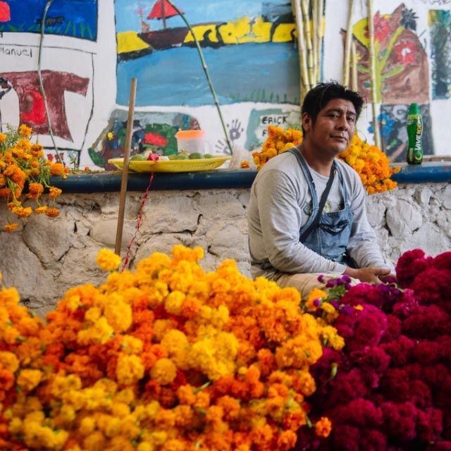 Man seated beside bright marigold and red flower piles at a colorful market stall