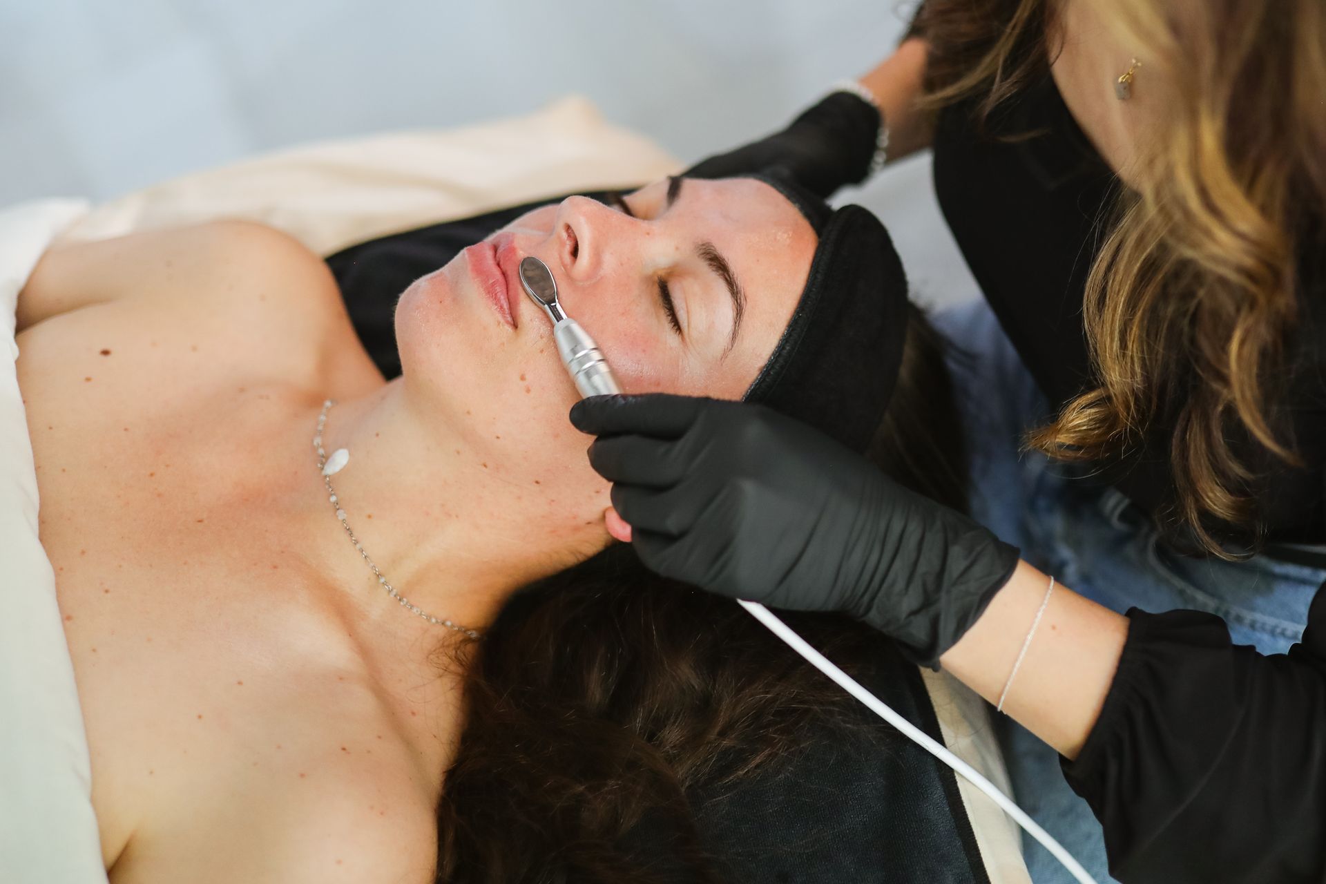 A person receives a professional facial treatment with an electronic device held by an aesthetician in black gloves.