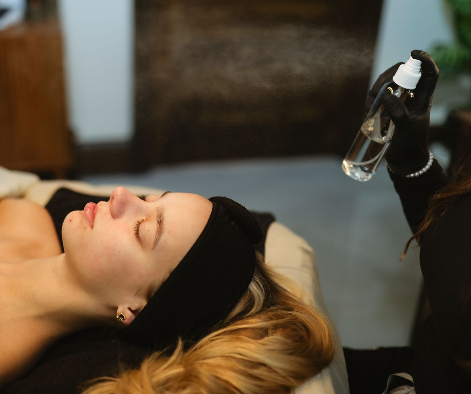 A woman is laying on a bed getting a spray on her face.