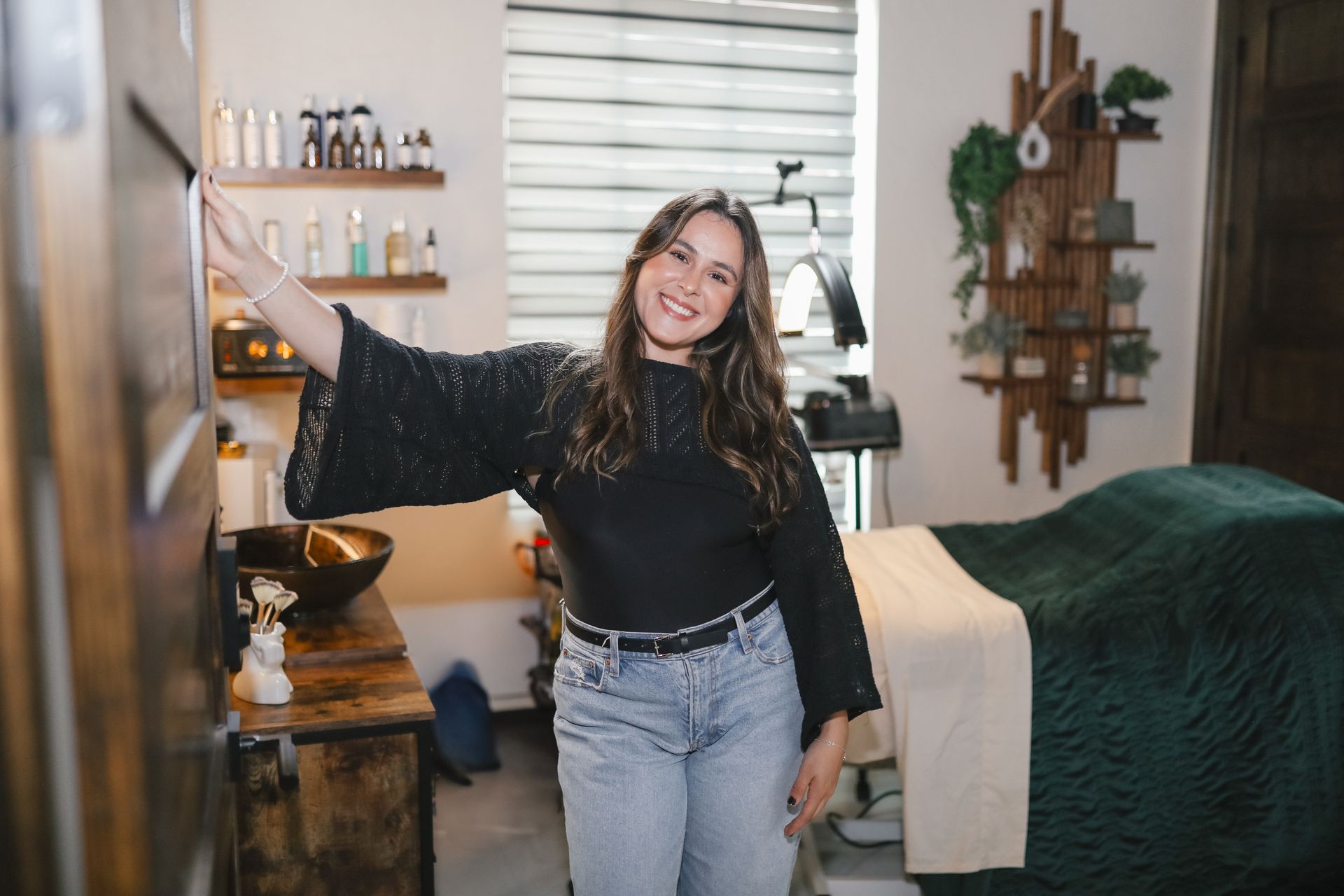 A smiling person in a black long-sleeved top and jeans standing in a treatment room with a massage table and wall shelving.