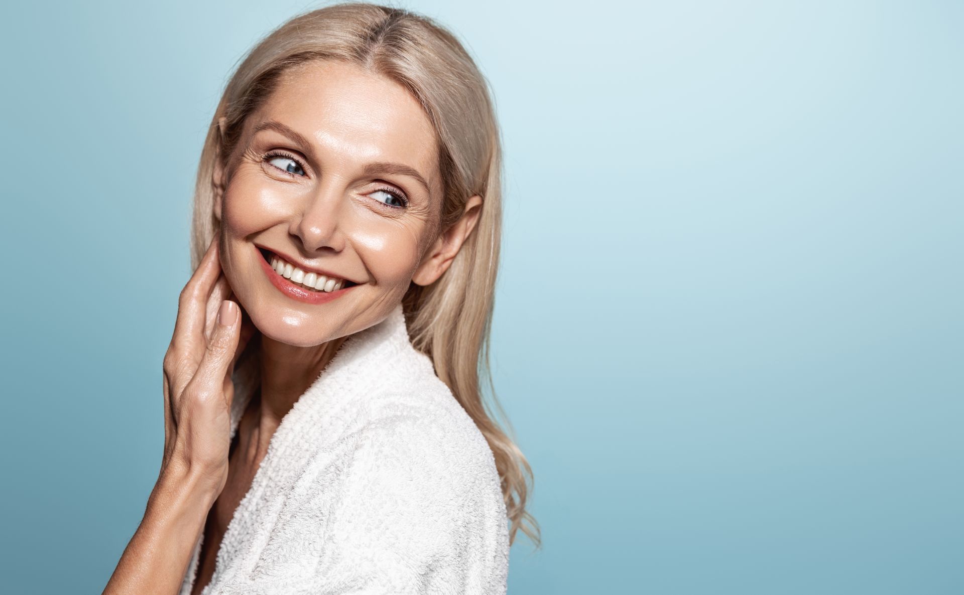 A smiling person with blonde hair wearing a white robe looks away against a soft blue background.