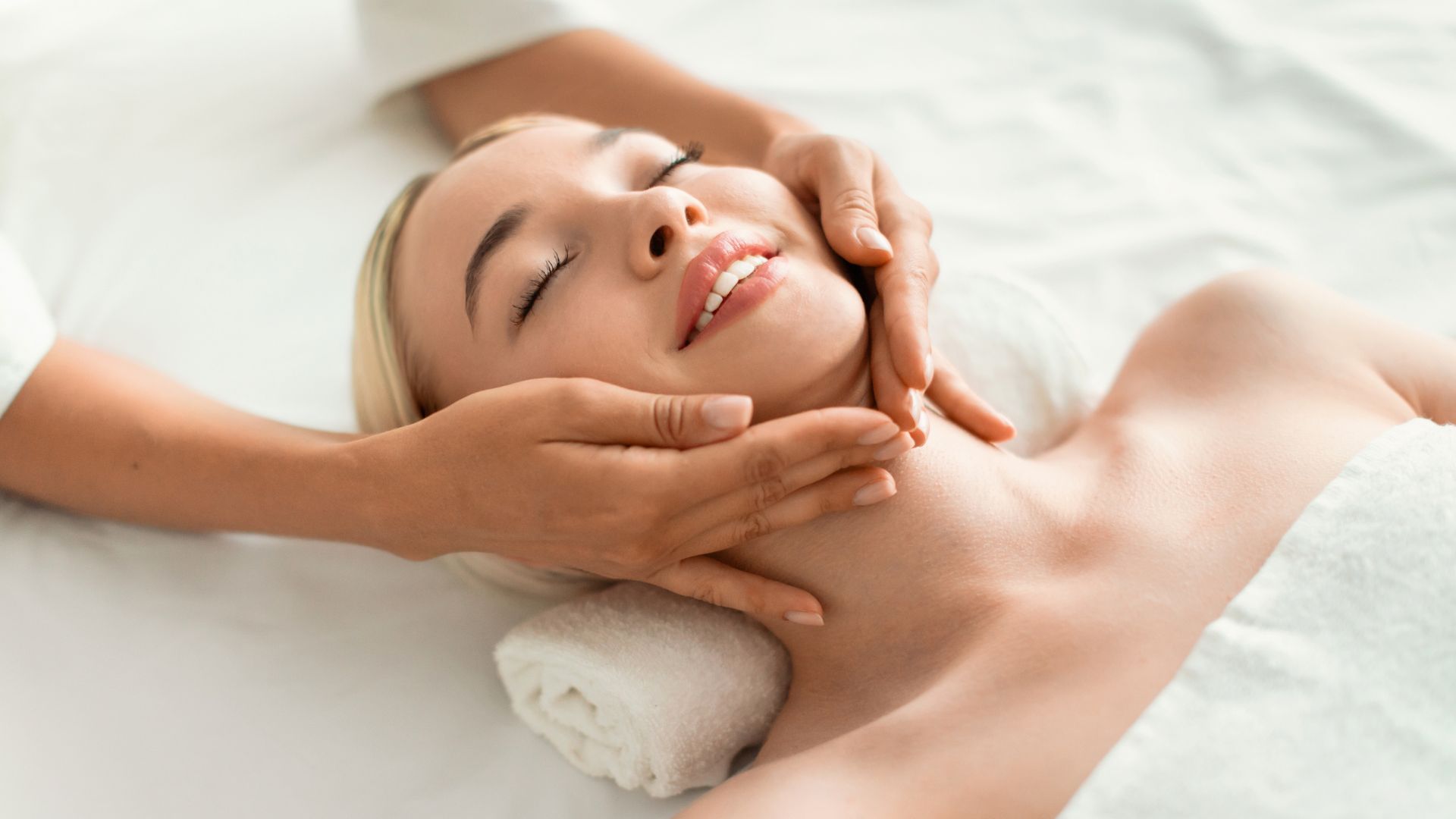 Person receiving a facial massage while lying on a white spa bed, smiling with eyes closed.