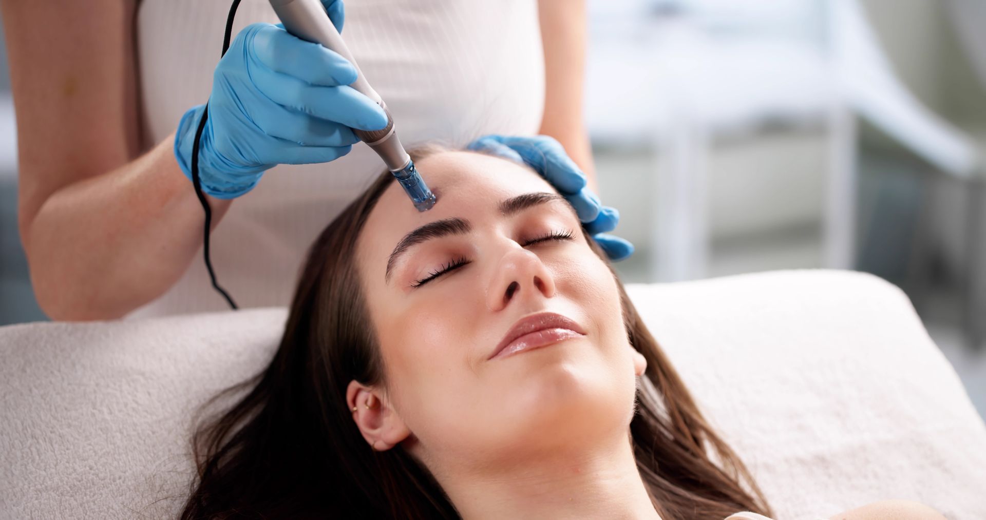 Woman receiving facial treatment, blue-gloved hands holding device to her forehead; spa setting.