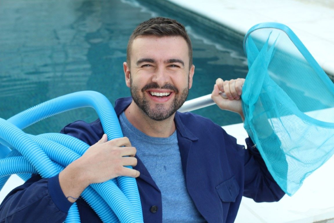 Man smiling by a pool, holding blue hoses and netting in a cleaning or maintenance setting