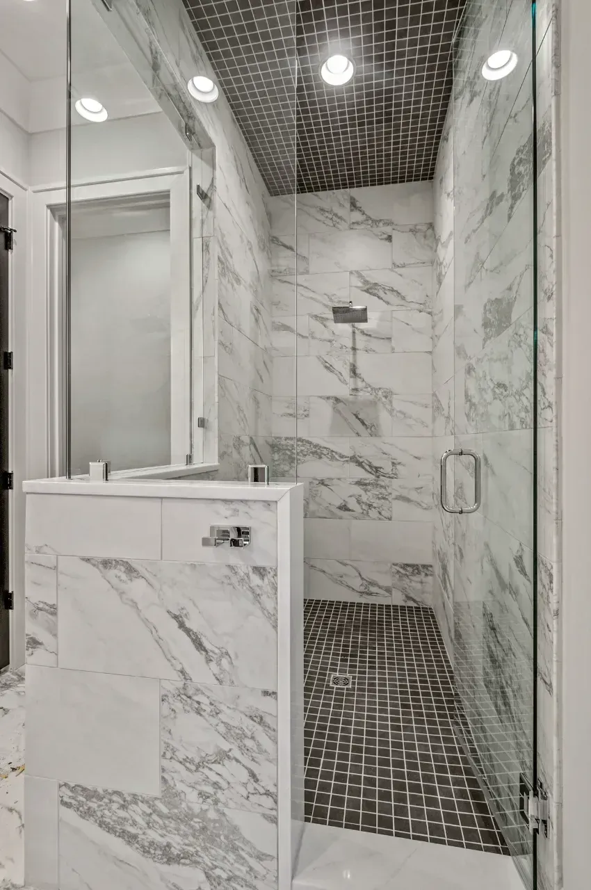 Modern marble-tiled shower with glass door, dark mosaic floor, and white vanity in a bathroom