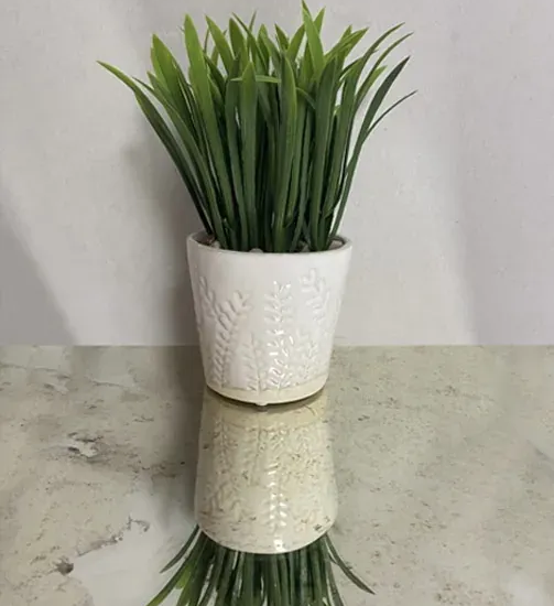 White textured pot with green artificial grass on a reflective tabletop.