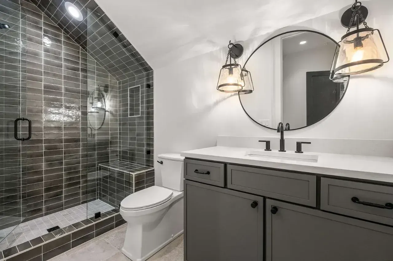 Modern bathroom with glass shower, toilet, gray vanity, round mirror, and brass light fixtures