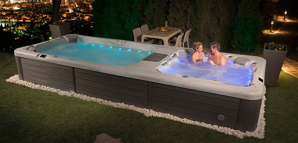 Two people relax in a hot tub on a grassy patio at night, with city lights in the background.