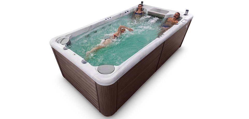 A woman swims in a long, rectangular hot tub. Two people relax in the back. The tub is brown with white trim.