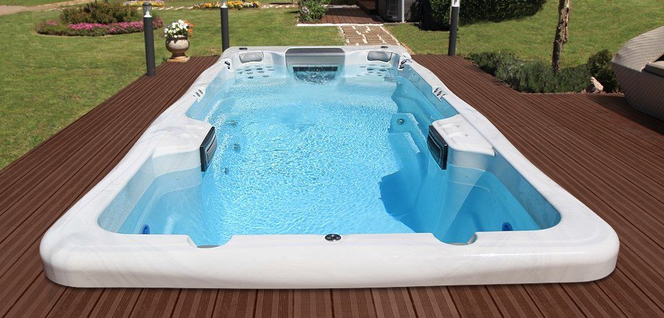 A white rectangular swim spa filled with blue water, set on a brown wooden deck outdoors.