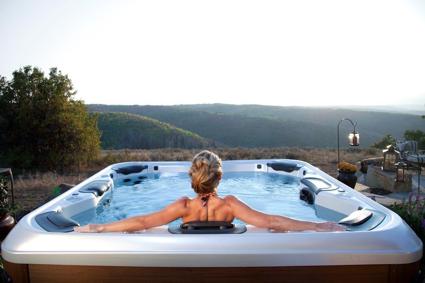Woman relaxes in a hot tub overlooking a scenic landscape at sunset.