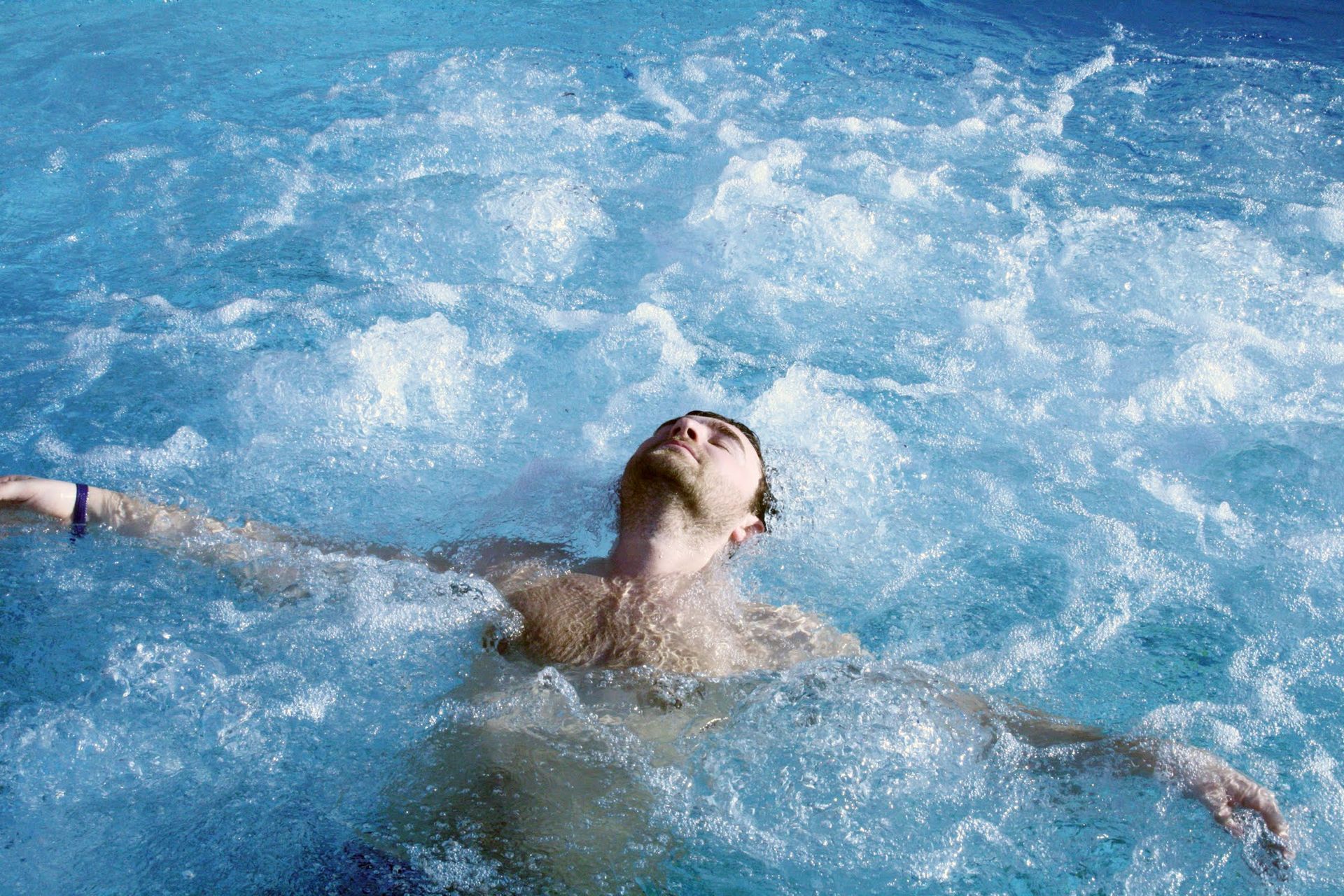 Man floating in blue water, arms outstretched, head tilted back, outdoors.