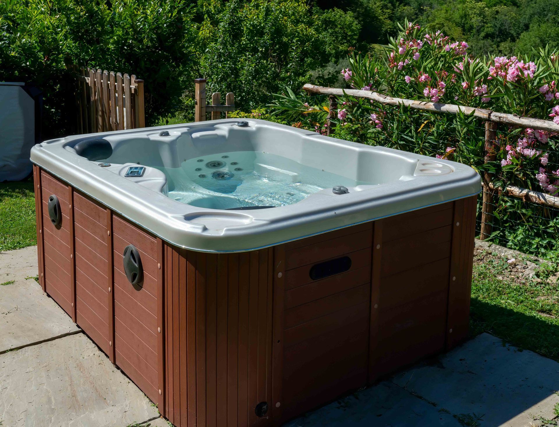 Outdoor hot tub with light blue water on a patio beside greenery and pink flowers