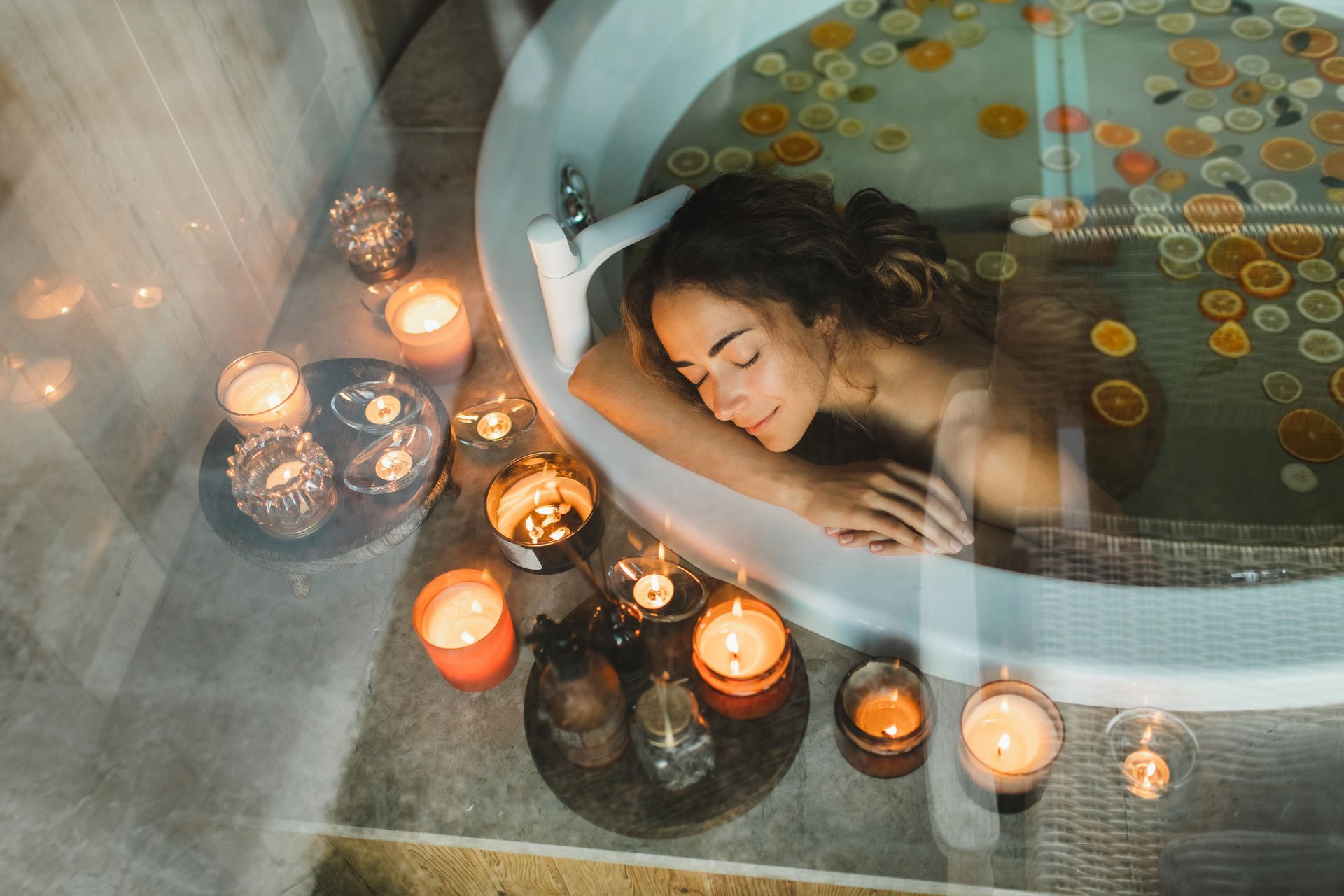 Woman relaxing in a bathtub with citrus slices, surrounded by lit candles, creating a spa-like atmosphere.
