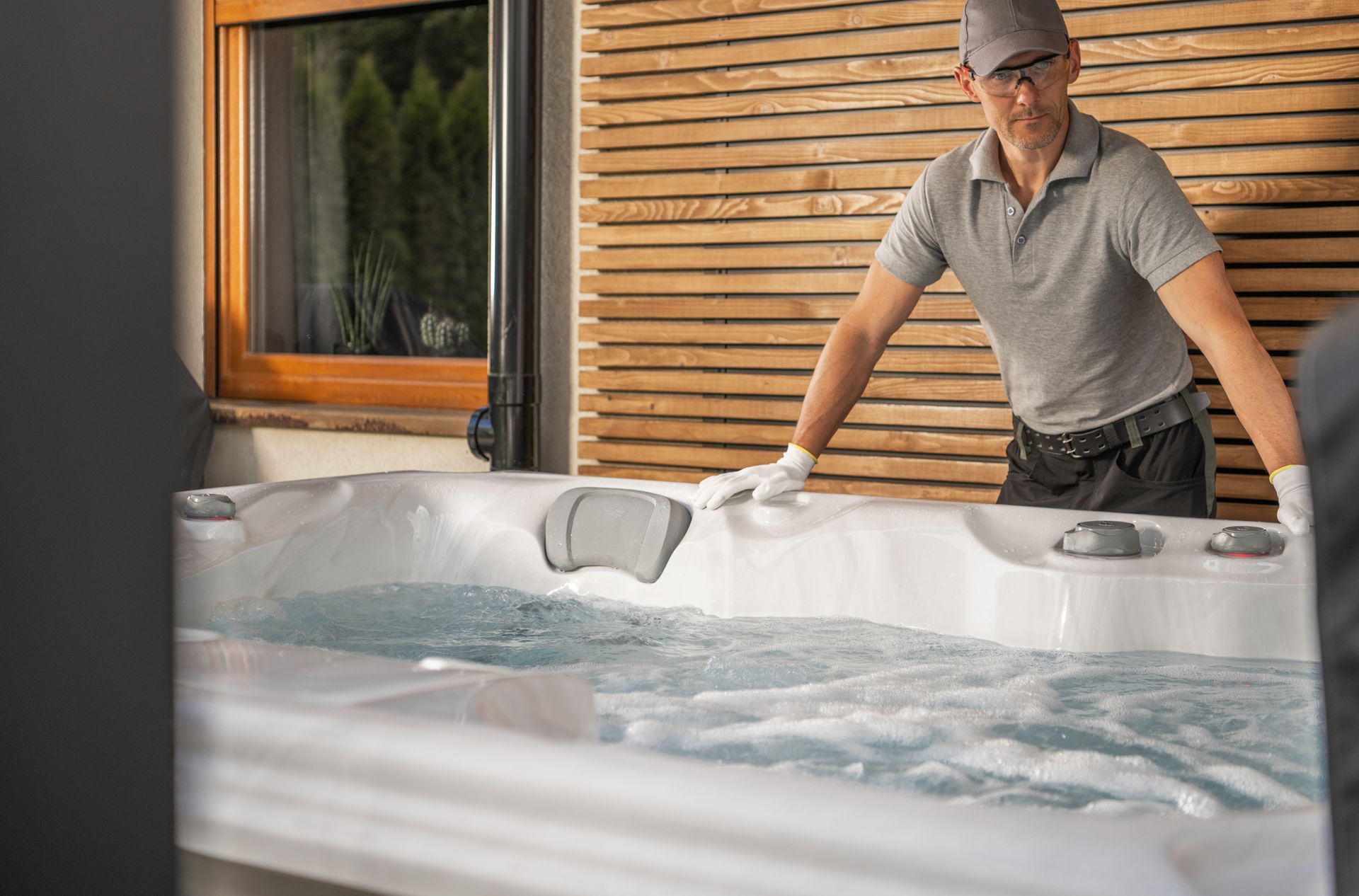 Man in gray shirt, gloves, and cap, inspecting a bubbling hot tub. Wooden wall background.