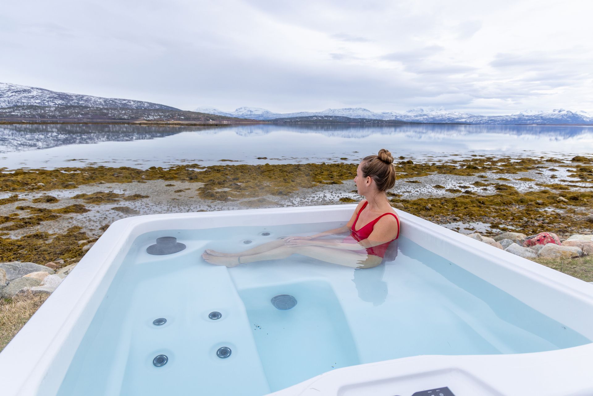 Woman in red swimsuit relaxes in hot tub by lake, mountains in background.
