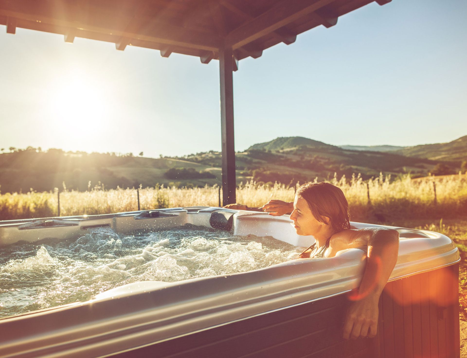 Woman relaxing in a hot tub outdoors, bathed in sunlight, with a scenic mountain view.
