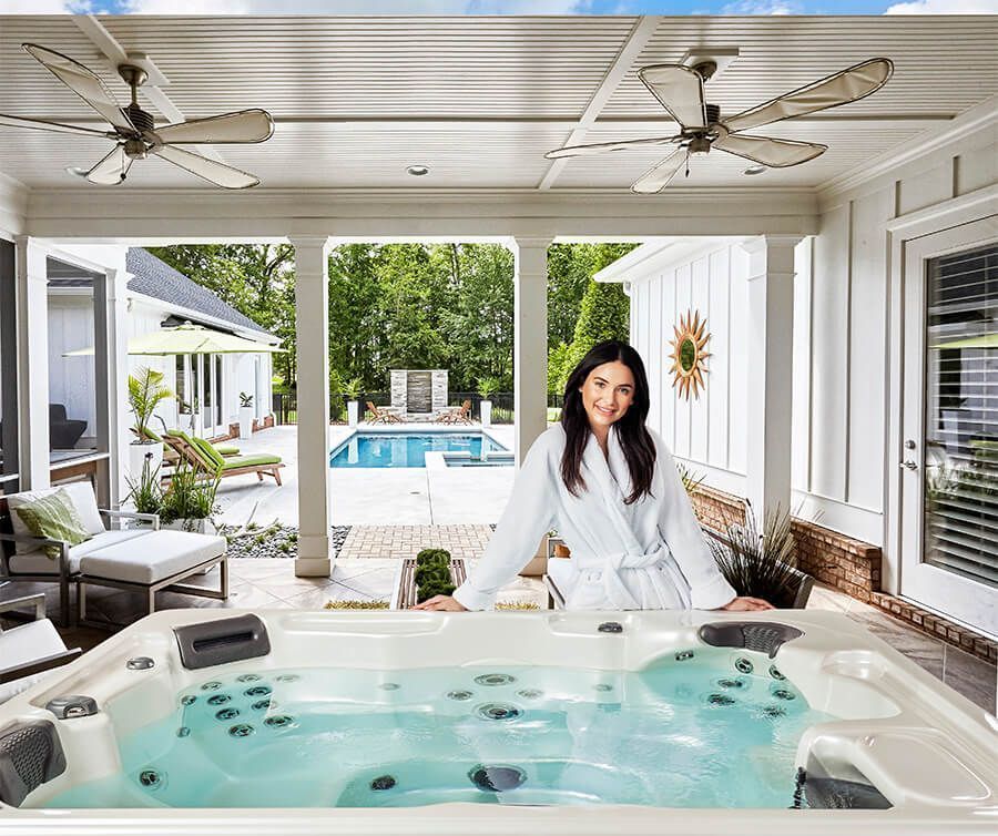 A person in a white bathrobe stands beside a lit hot tub on a covered patio overlooking a sunny swimming pool and garden.