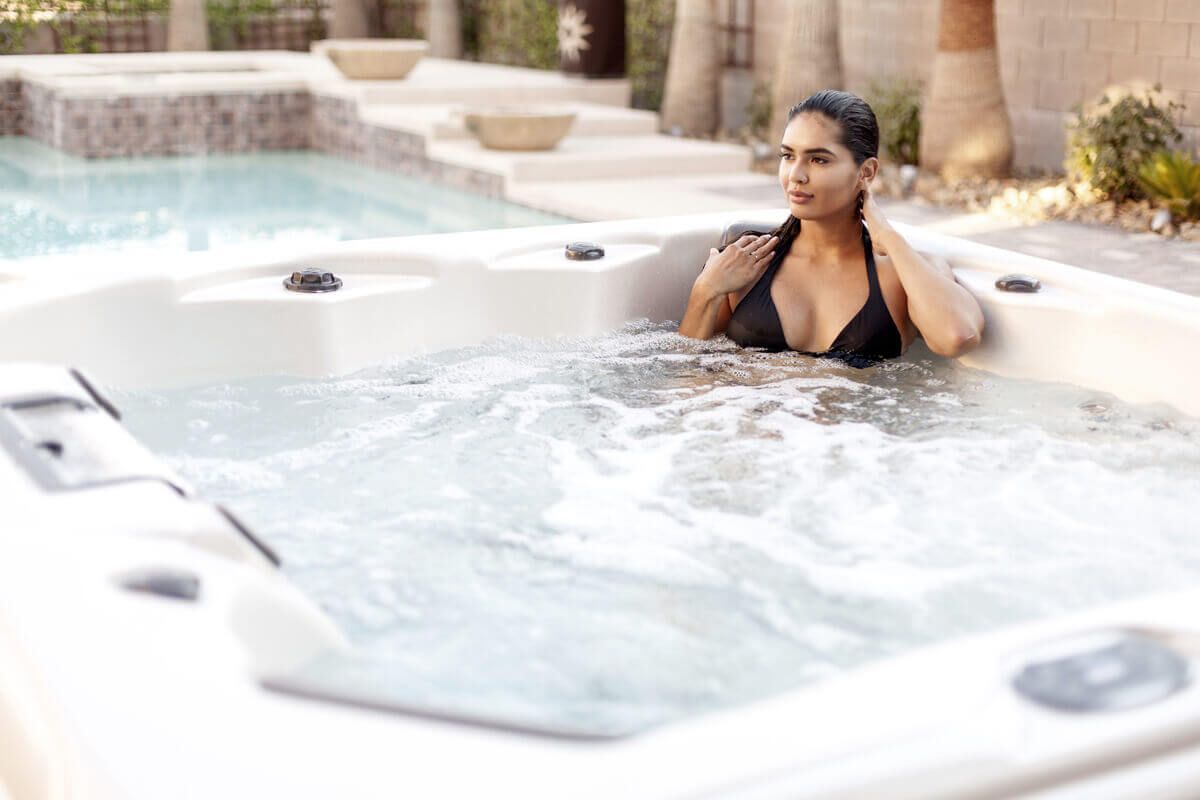 A person relaxes in a bubbling outdoor hot tub with a swimming pool and patio in the background.