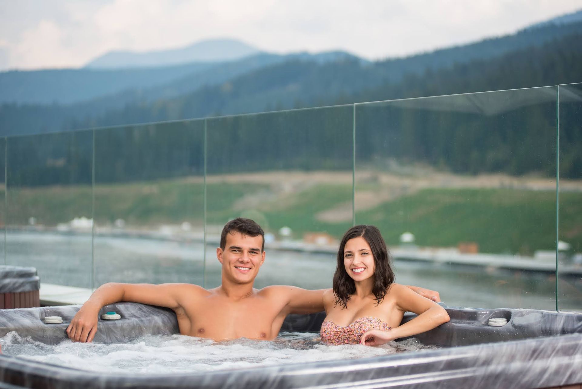 Couple smiling in a hot tub, overlooking a scenic mountain landscape and lake.