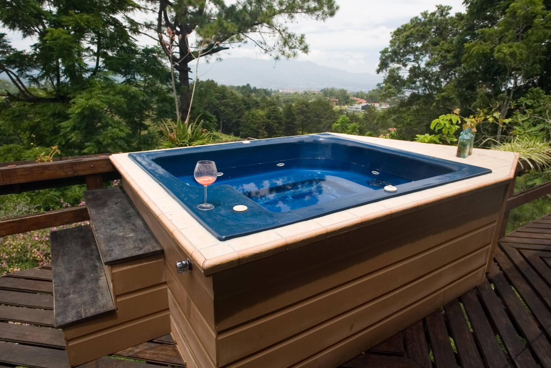 Hot tub on deck overlooking lush green landscape with drink.