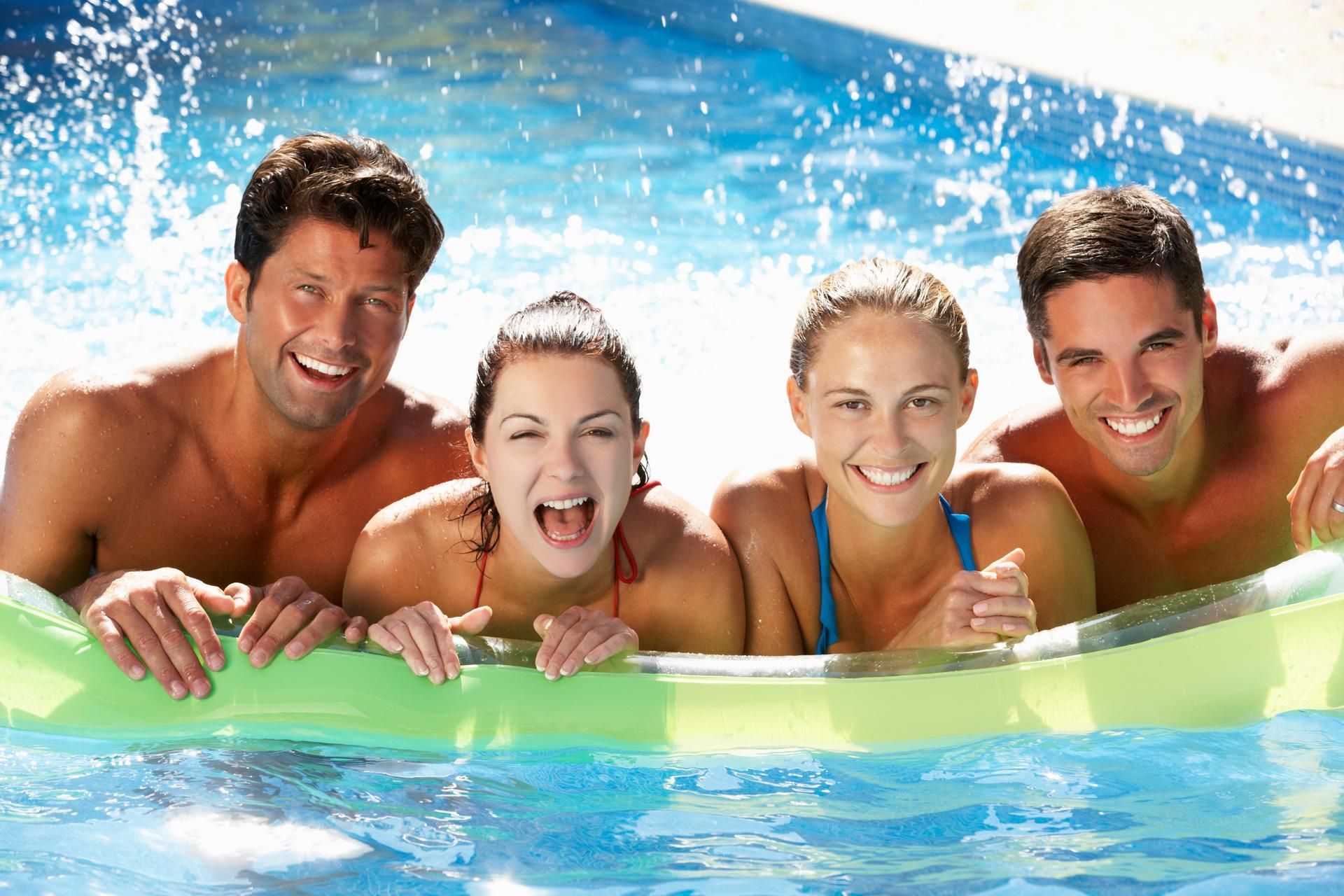 Four smiling people in a pool on a raft. Splashing water and sunny day.