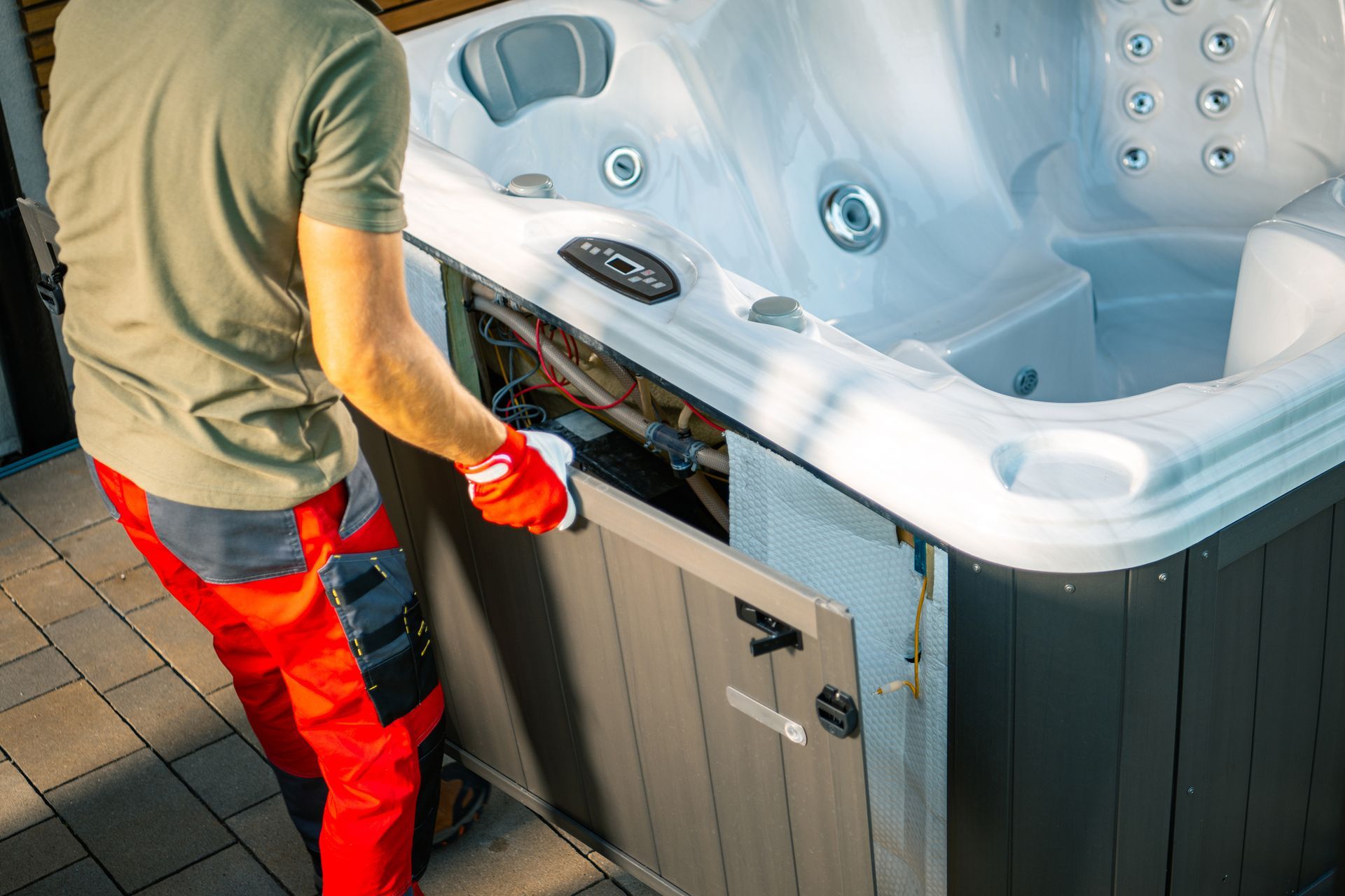 Man in red pants repairs hot tub panel, exposing internal components.