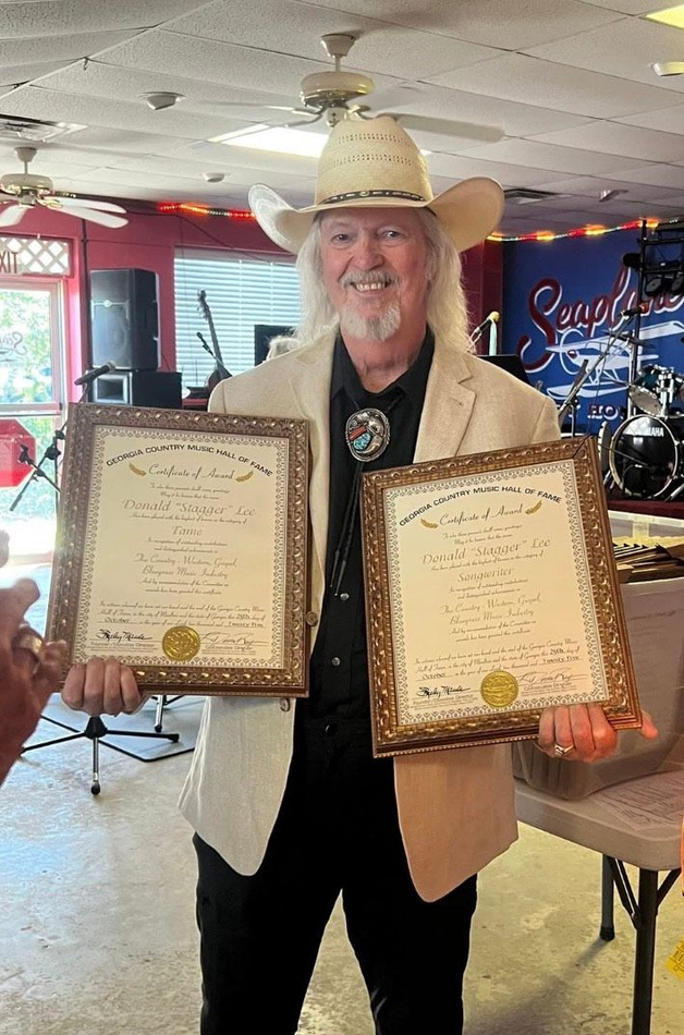 Man in cowboy hat holding two framed certificates inside a venue.