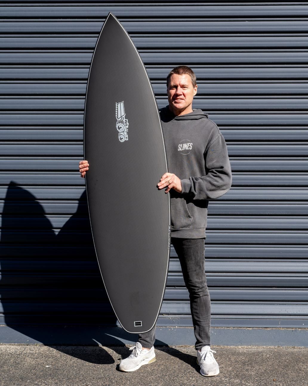A Man Is Holding a Black Surfboard in Front of A Garage Door — Slimes Boardstore in Bateau Bay, NSW