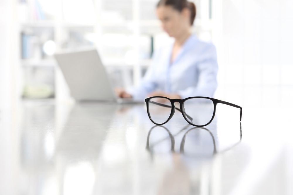 A Woman Is Sitting at A Desk with A Laptop and Glasses on It — John Williams Optometrist in Grafton, NSW