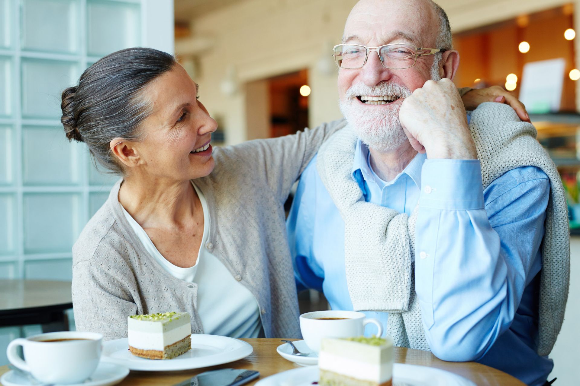 Two people smiling at a café table with coffee cups and dessert, one leaning toward the other
