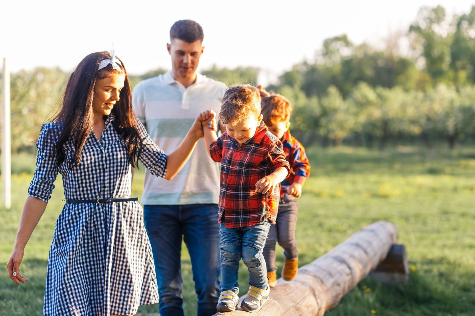 Family walking along a log in a sunny grassy field, with an adult holding a child’s hand.