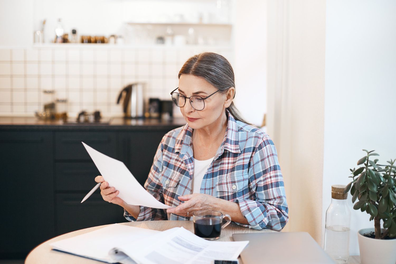 Person reading papers at a kitchen table with a laptop, coffee cup, and houseplant nearby